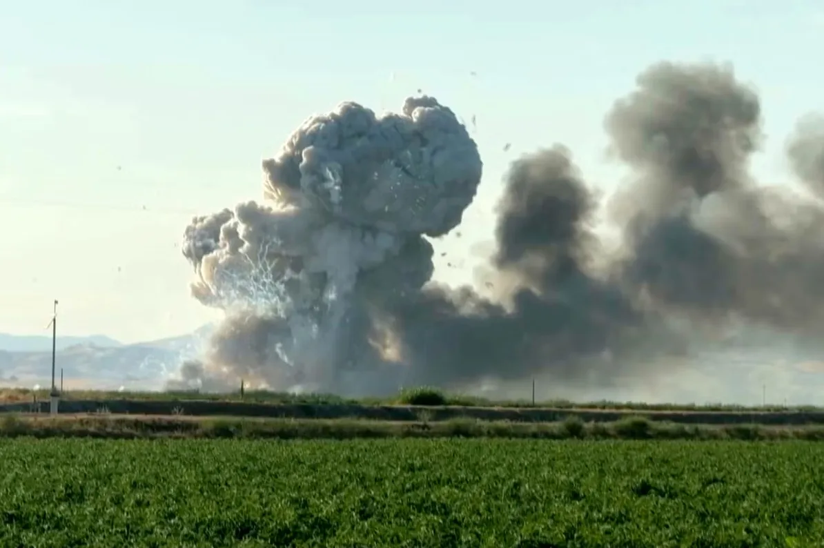 Velika eksplozija sa gustim dimom i krhotinama na nebu, iznad zelenog polja i brdovitog pejzaža.