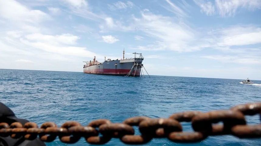 Veliki teretni brod usidren na pučini, sa lancima spuštenim u vodu i manjim patrolnim plovilom koje prolazi u daljini.