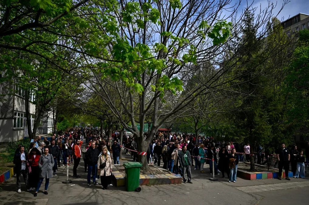Grupa ljudi stoji u dugom redu na otvorenom, pored zgrada i drveća prekrivenih zelenilom.