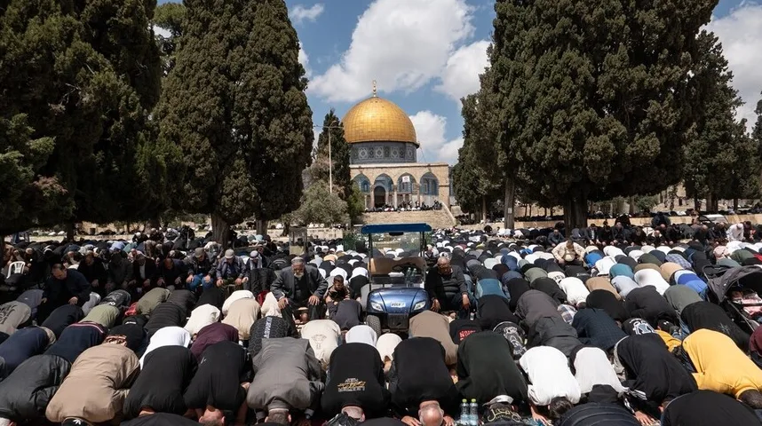 Veliki broj muškaraca u bijelim i tamnim odorama na koljenima u molitvi ispred Kupole na stijeni, sa drvećem u prvom planu i vedrim nebom.