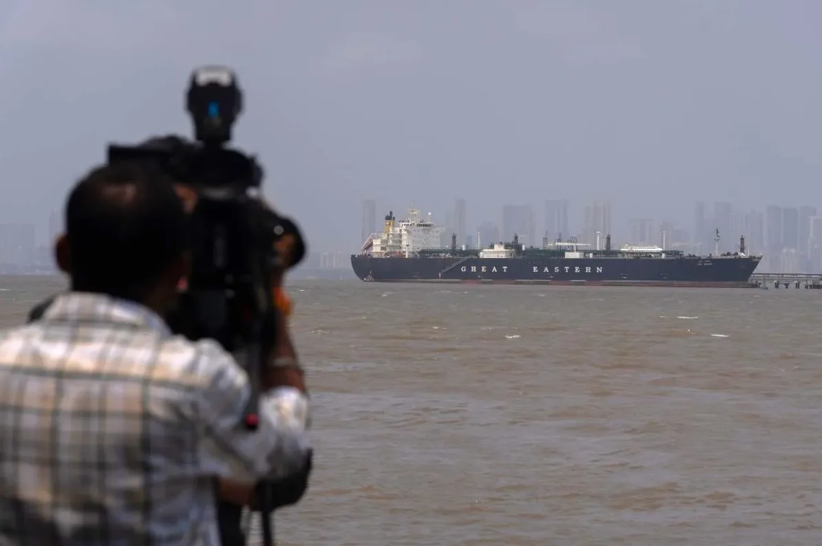 Krupni plan kamermana koji snima veliki teretni brod nazvan "Great Eastern" kako plovi prema obali s gradskim neboderima u pozadini.