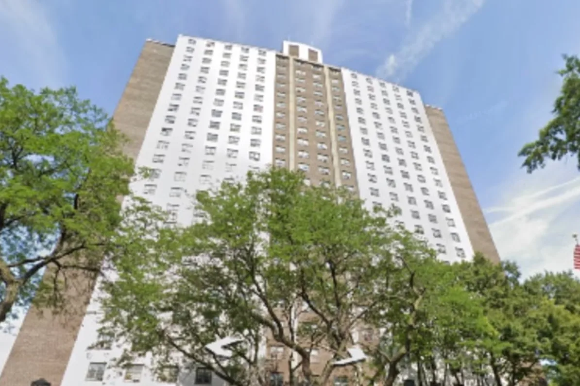 Visoki stambeni objekat sa bijelom fasadom i smeđim centralnim dijelom, okružen zelenim drvećem pod vedrim plavim nebom.