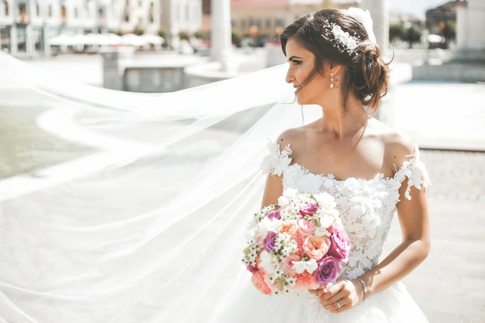 Vjenčana mlada u raskošnoj bijeloj vjenčanici sa velom koji leprša, drži buket raznobojnog cvijeća i gleda u daljinu.