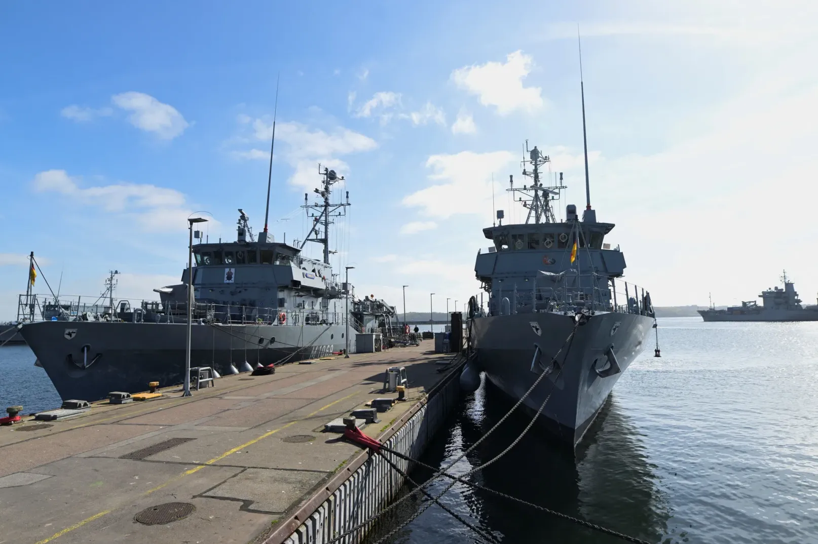 Dva siva vojna broda usidrena jedan pored drugog u luci na sunčan dan.