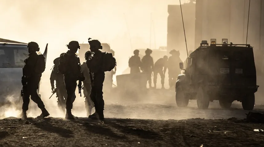 Siluete vojnika u vojnoj opremi i s oružjem, dok se iza njih vidi oklopno vozilo i zgrade u magli.