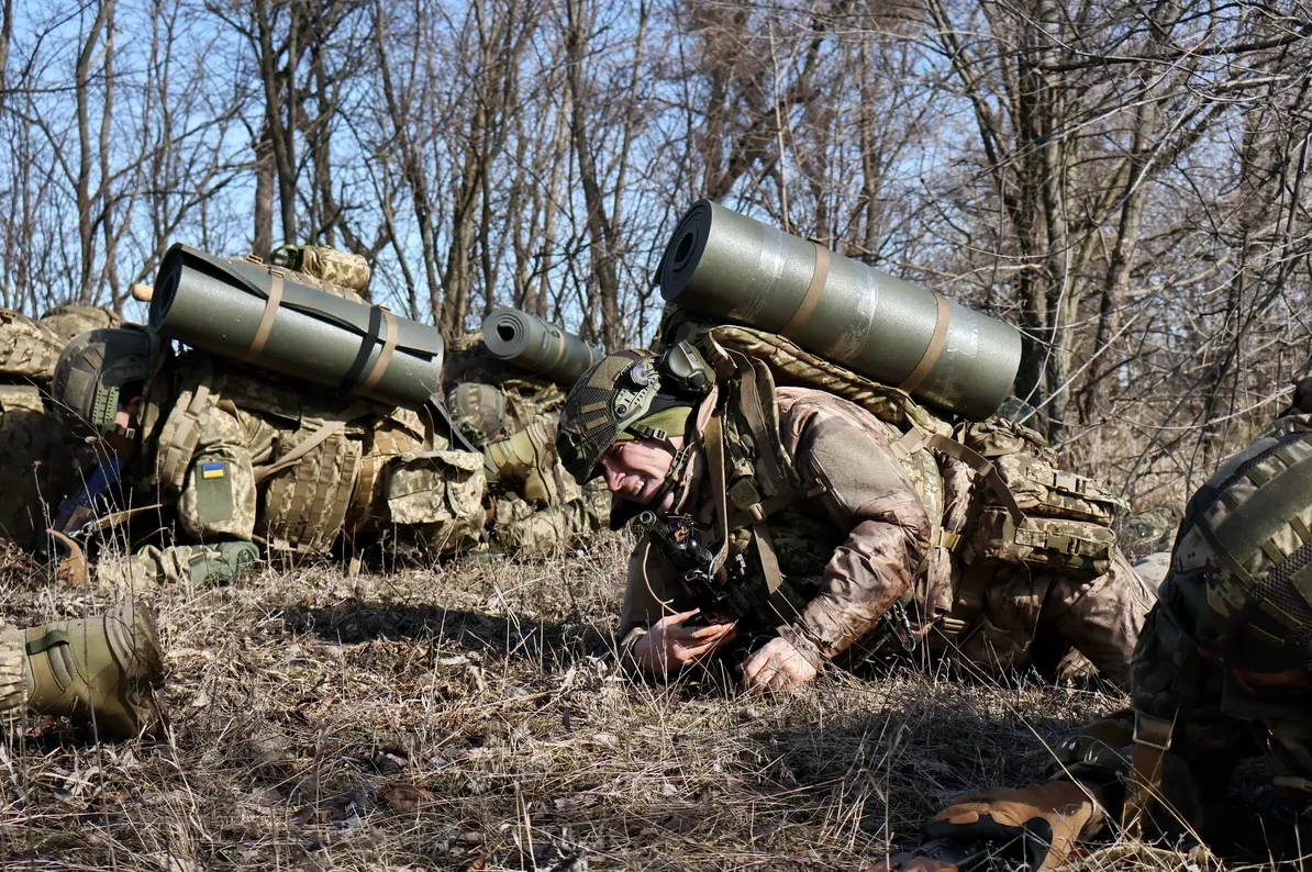 Ukrajinski vojnici opremljeni rančevima i vojnom opremom, leže u travi pored drveća.