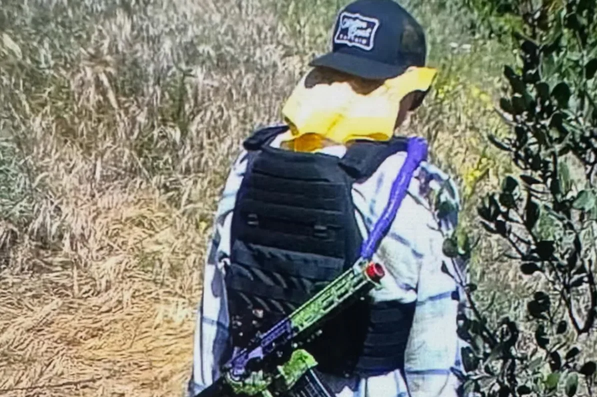 Osoba u vojnom prsluku i kačketu, sa žutom maramom oko lica i jarkozelenom puškom.