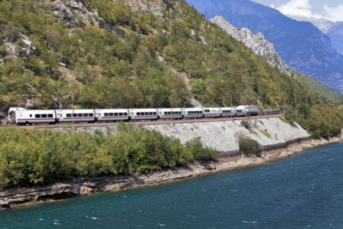 Bijeli putnički voz se kreće lijevom stranom pruge uz planinu prekrivenu drvećem, tik uz tirkiznu rijeku.