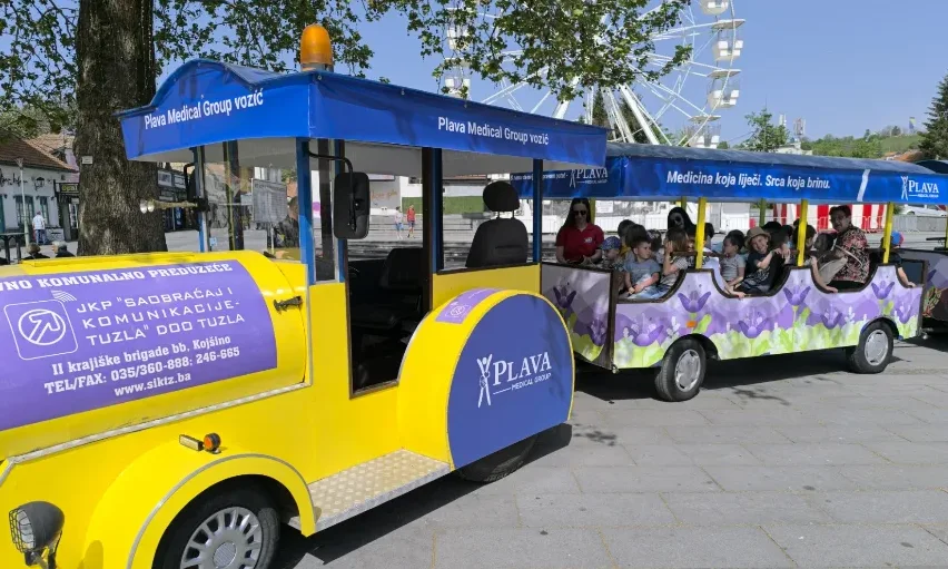 Žuti turistički vozić sa plavom nadstrešnicom, pun djece, parkiran na popločanoj stazi. Na nadstrešnici piše "Plava Medical Group vozić". Na jednom od vagona su ljubičasti cvjetovi.