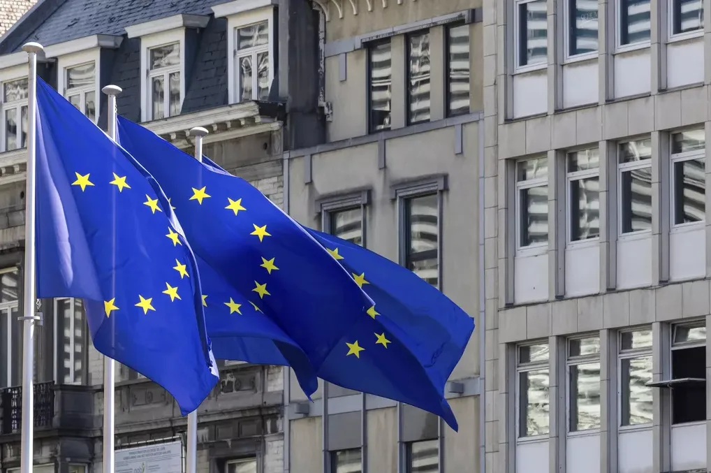 Dvije zastave Evropske unije vijore se na vjetru ispred modernih zgrada sa mnogo prozora.