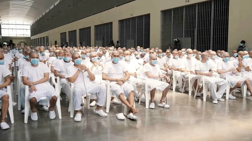 Veliki broj zatvorenika u bijelim majicama i kratkim hlačama sjedi na bijelim stolicama u zatvorskoj ćeliji, svi nose plave hirurške maske.