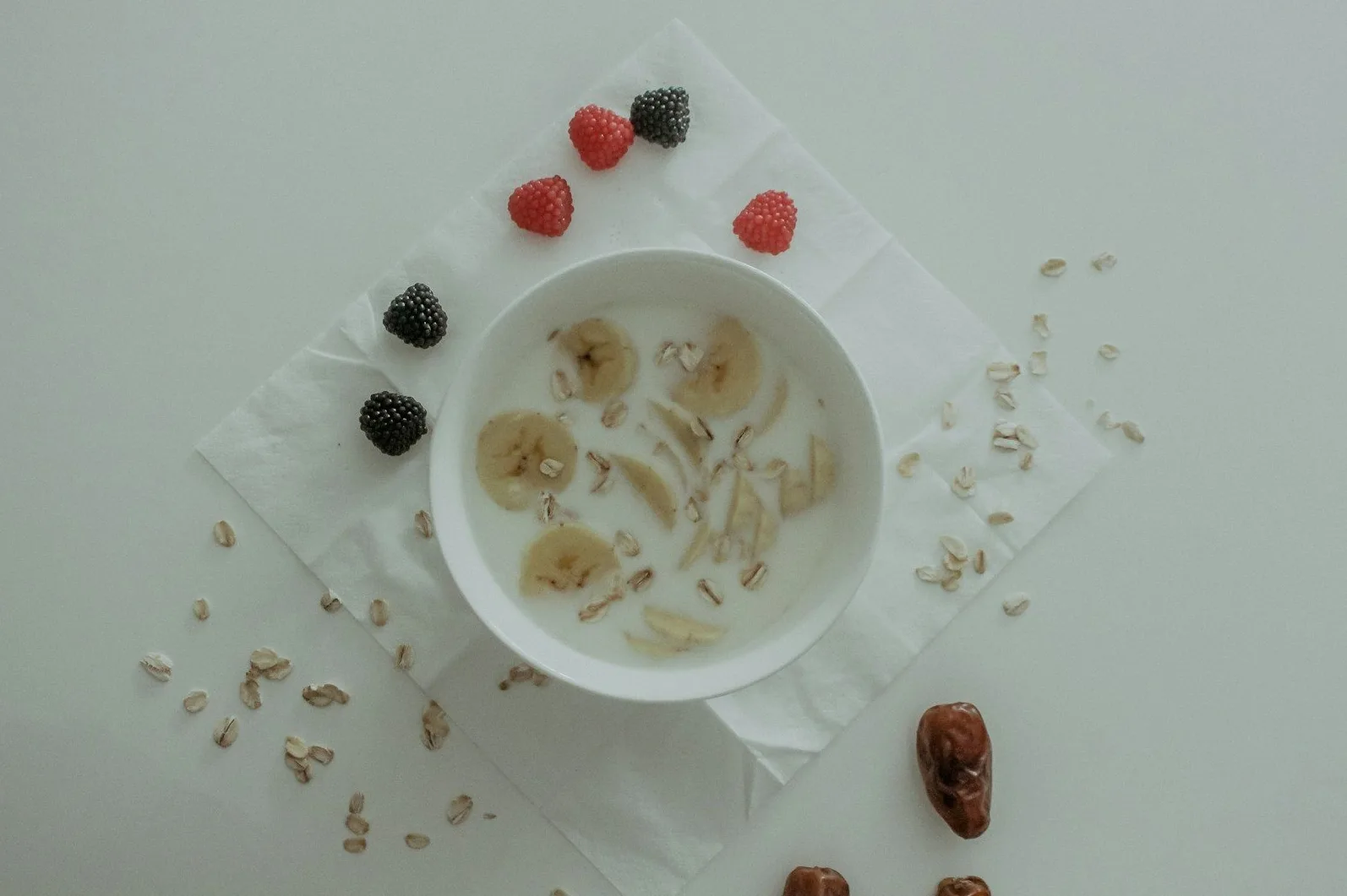 Zdjela sa zobenim pahuljicama, kriškama banane i mlijekom, ukrašena kupinama i malinama, s dodatnim zobenim pahuljicama i urmom raspoređenim po stolu.