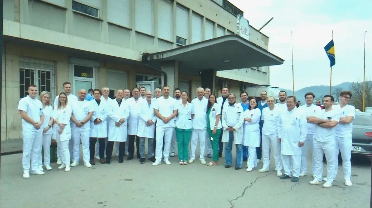 Grupa zdravstvenih radnika u bijelim uniformama pozira ispred bolnice sa zastavom Bosne i Hercegovine.