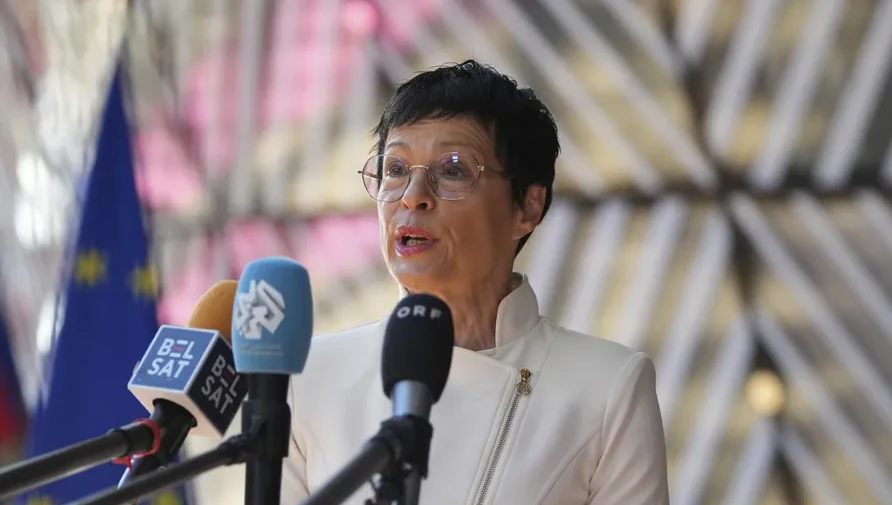 Žena u bijelom sakou sa naočarima govori ispred više mikrofona.