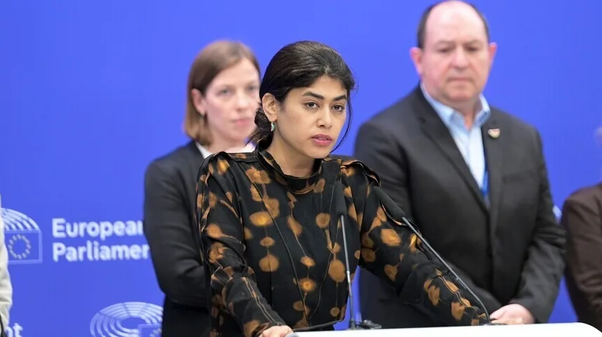 Žena sa tamnom kosom, noseći crnu haljinu sa zlatnim dezenom, govori iza podijuma. U pozadini se vide dva lica, jedno žensko i jedno muško, a na lijevoj strani se nazire logo Evropskog parlamenta.