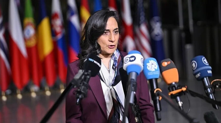 Žena u ljubičastom sakou i maramom oko vrata govori pred brojnim mikrofonima. U pozadini su vidljive brojne zastave različitih država.