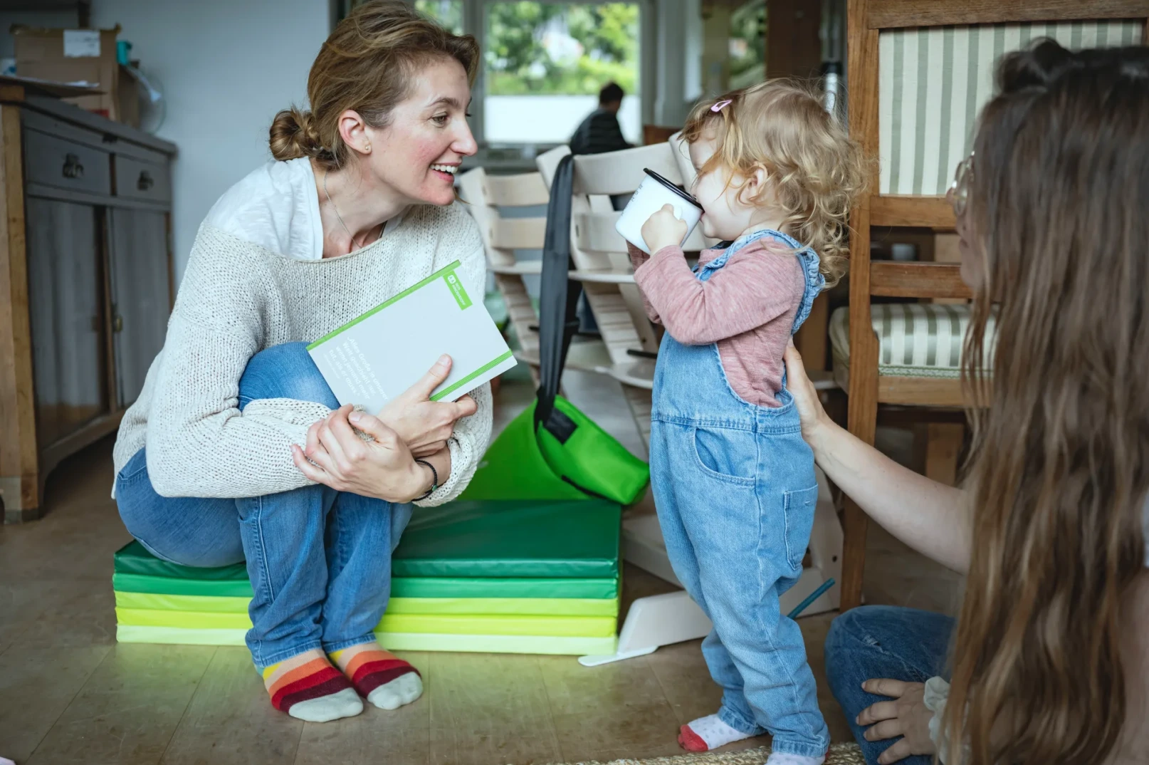 Žena sjedi na podu držeći knjigu i gleda u malo dijete koje pije iz šolje.
