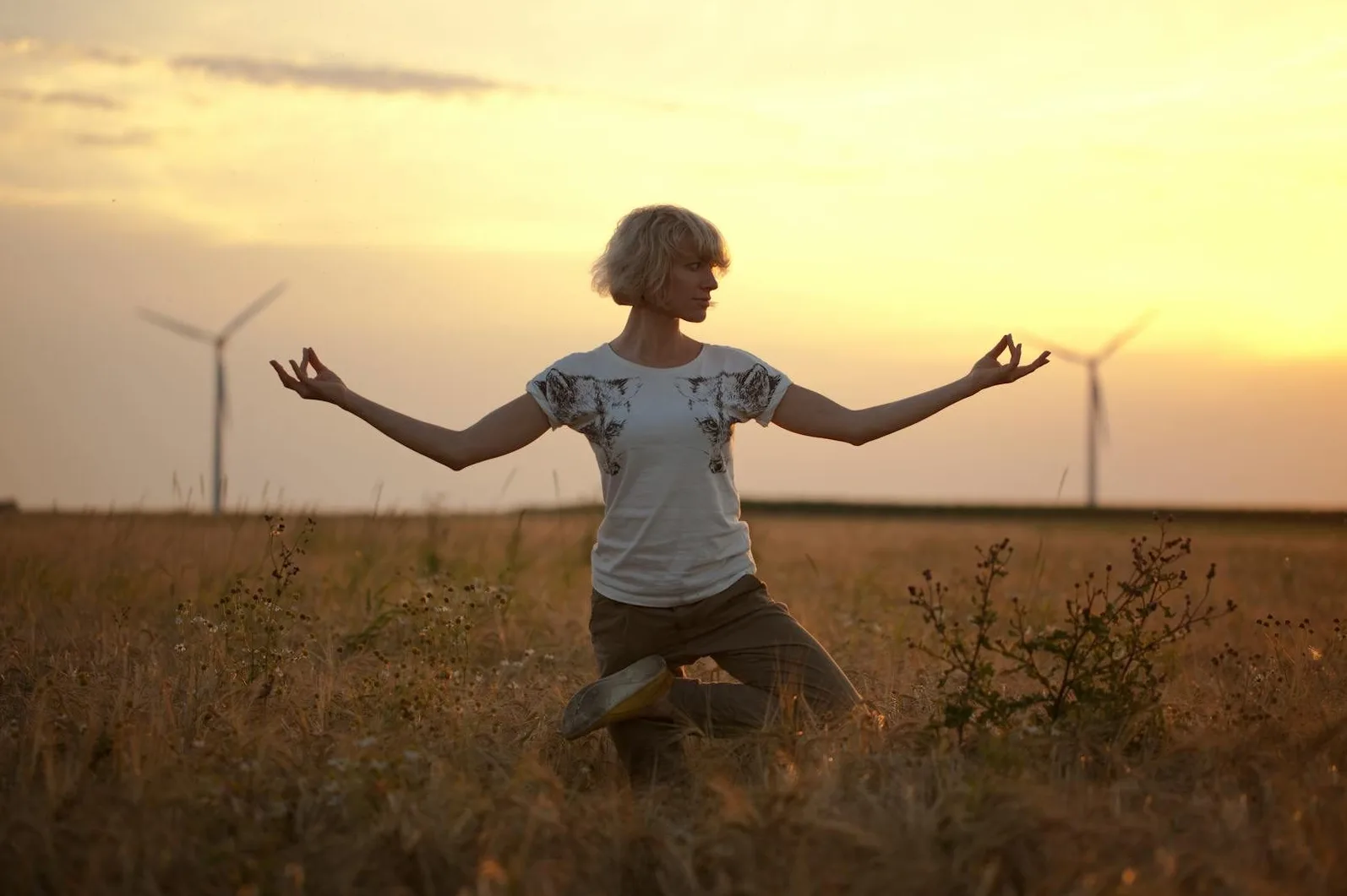 Žena izvodi vježbu joge u polju zlatnog žita sa vjetroelektranama u pozadini tokom zalaska sunca.