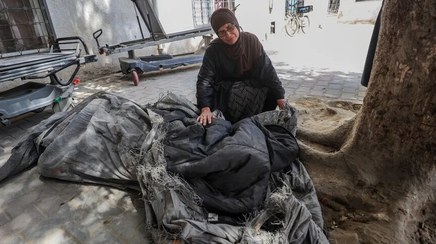 Žena u hidžabu i naočalama sjedi pored istrošenog pokrivača na trotoaru, sa korijenjem drveta u prvom planu.