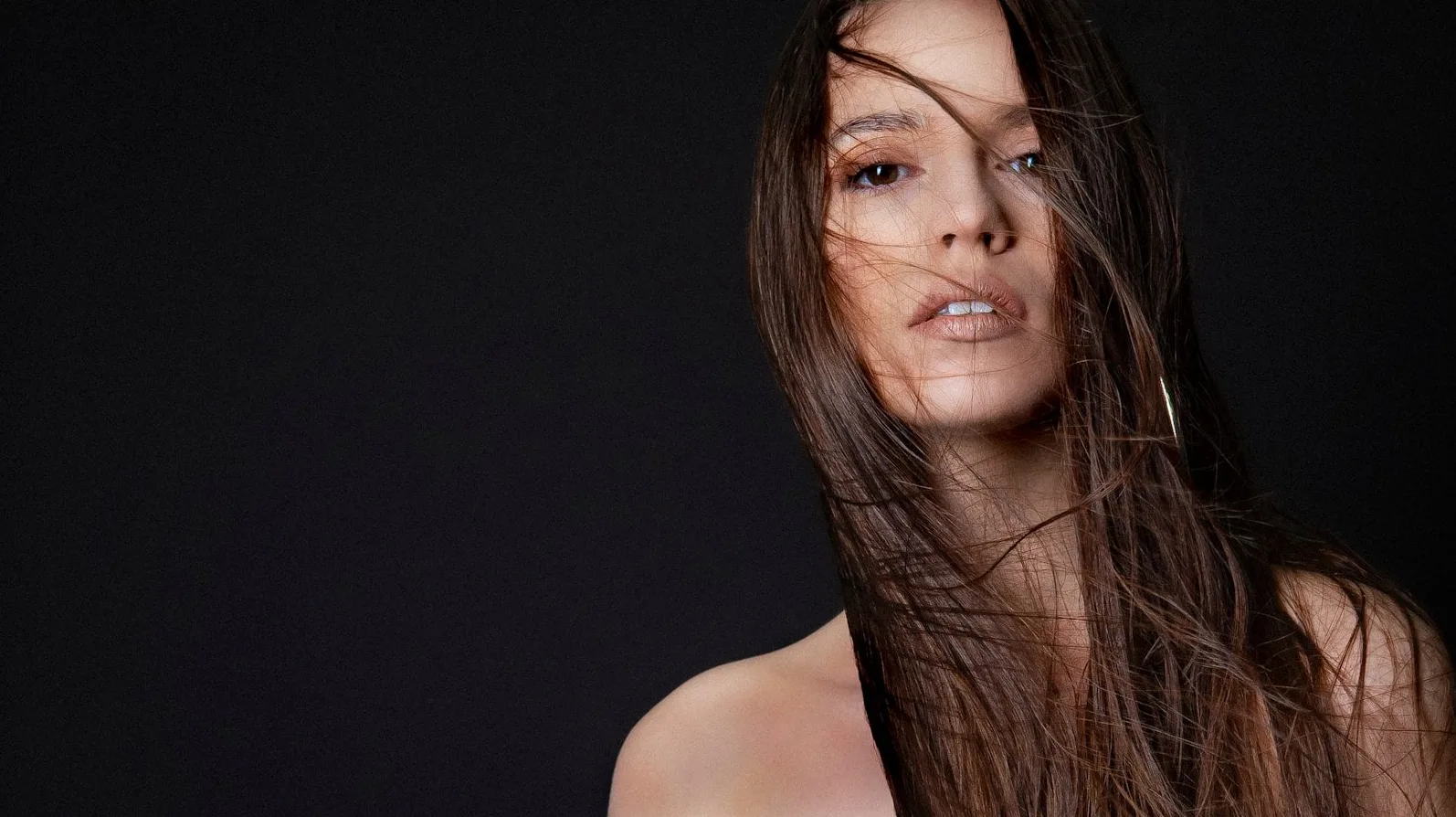 Portret mlade žene sa dugom, tamnom, talasastom kosom koja joj pada preko lica, na tamnoj pozadini.