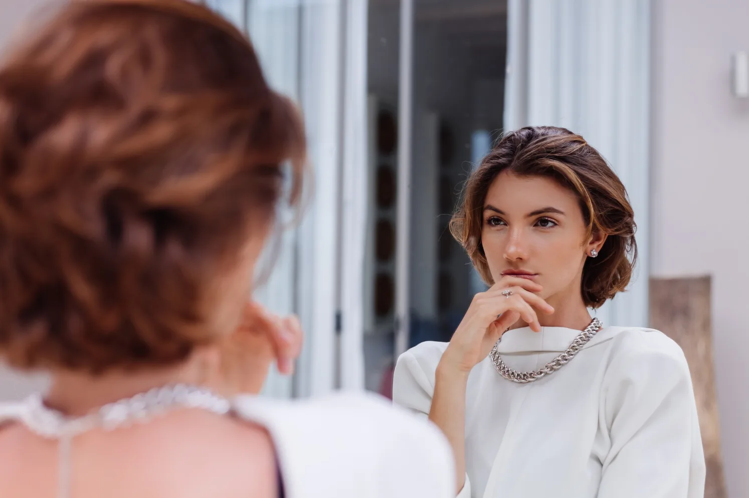 Mlada žena u bijeloj bluzi sa modernom ogrlicom gleda u ogledalo, sa rukom na obrazu.