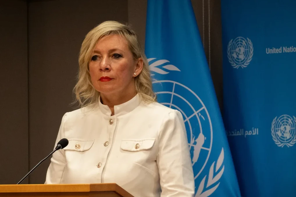 Žena sa plavom kosom i crvenim ružem, obučena u bijelo odijelo, stoji iza govorničkog postolja ispred plave zastave Ujedinjenih naroda.