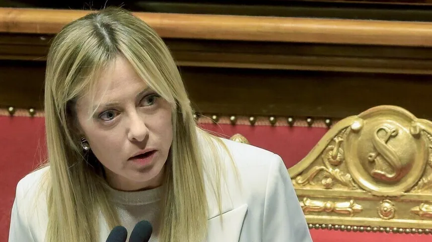 Žena u bijelom odijelu s plavom kosom govori ispred mikrofona u bogato uređenoj sali.