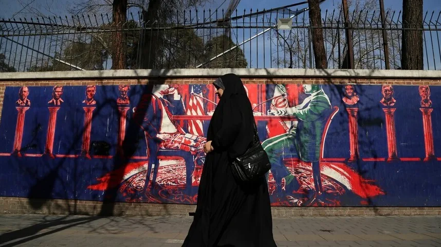 Žena u crnom hidžabu i dugoj haljini prolazi pored zida sa muralom na kojem su prikazane biste američkih predsjednika i scene koje simbolizuju politiku SAD-a i Irana.