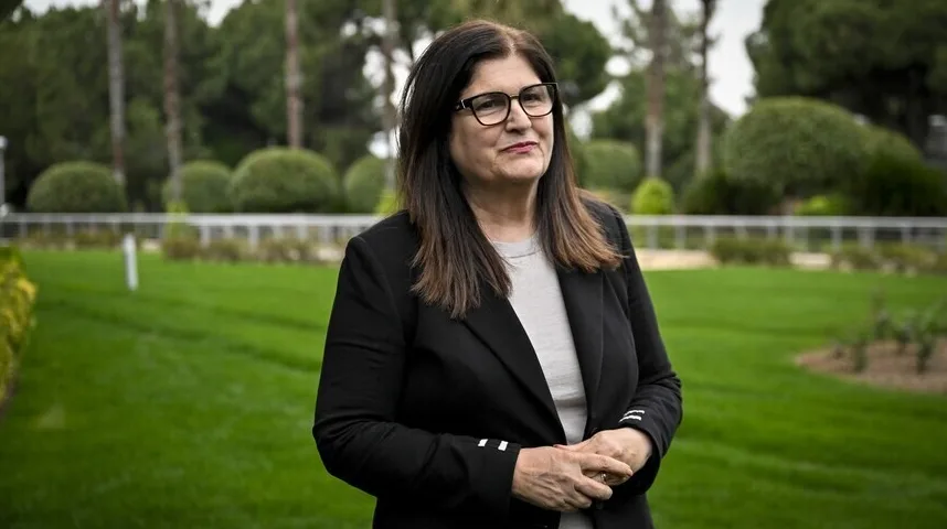 Žena sa smeđom kosom i naočalama nosi crni sako i sivu majicu dok stoji vani u parku.