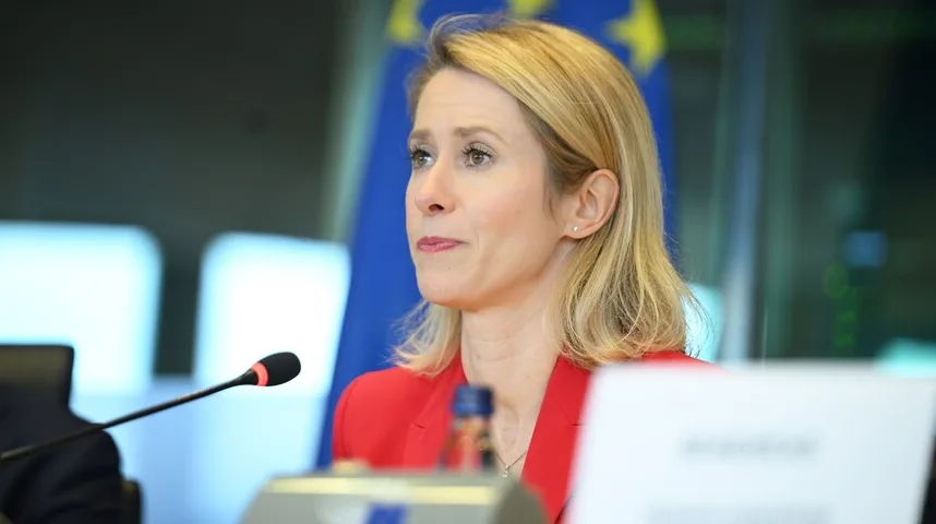 Plavokosa žena u crvenom sakou gleda lijevo, dok se ispred nje nalazi mikrofon i plava zastava sa žutim zvijezdama.