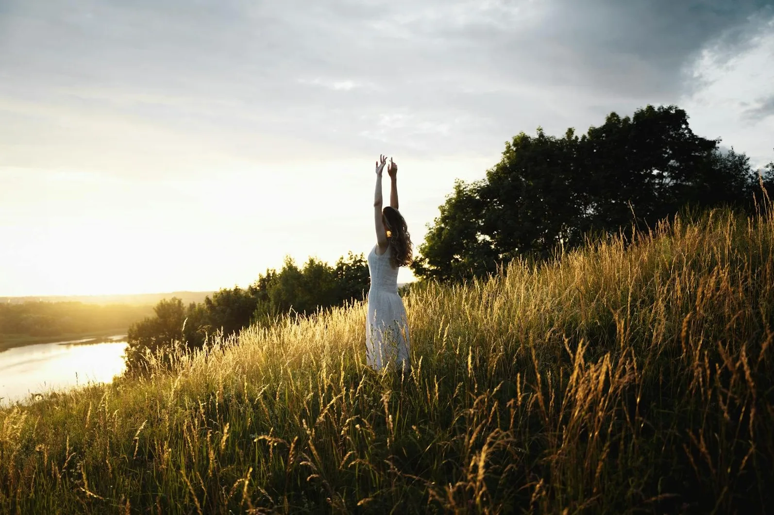 Žena u bijeloj haljini podiže ruke ka nebu, stojeći usred visokog, zlatnog žita na obronku brda, dok sunce zalazi iza drvoreda u daljini.