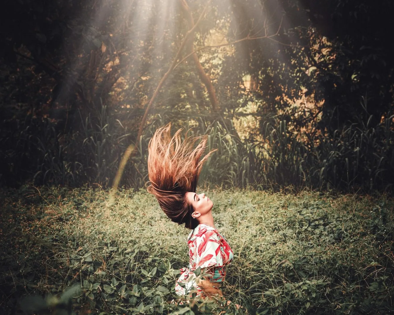 Žena sa dugom smeđom kosom koja se njiše u vazduhu, u cvjetnoj haljini, na pozadini bujne zelene vegetacije i sunčevih zraka koji prolaze kroz drveće.