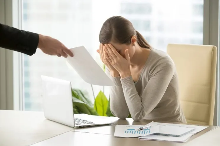 Tužna žena uredu drži ruke na licu, frustrirana ili razočarana dok joj neko dodaje dokumente.