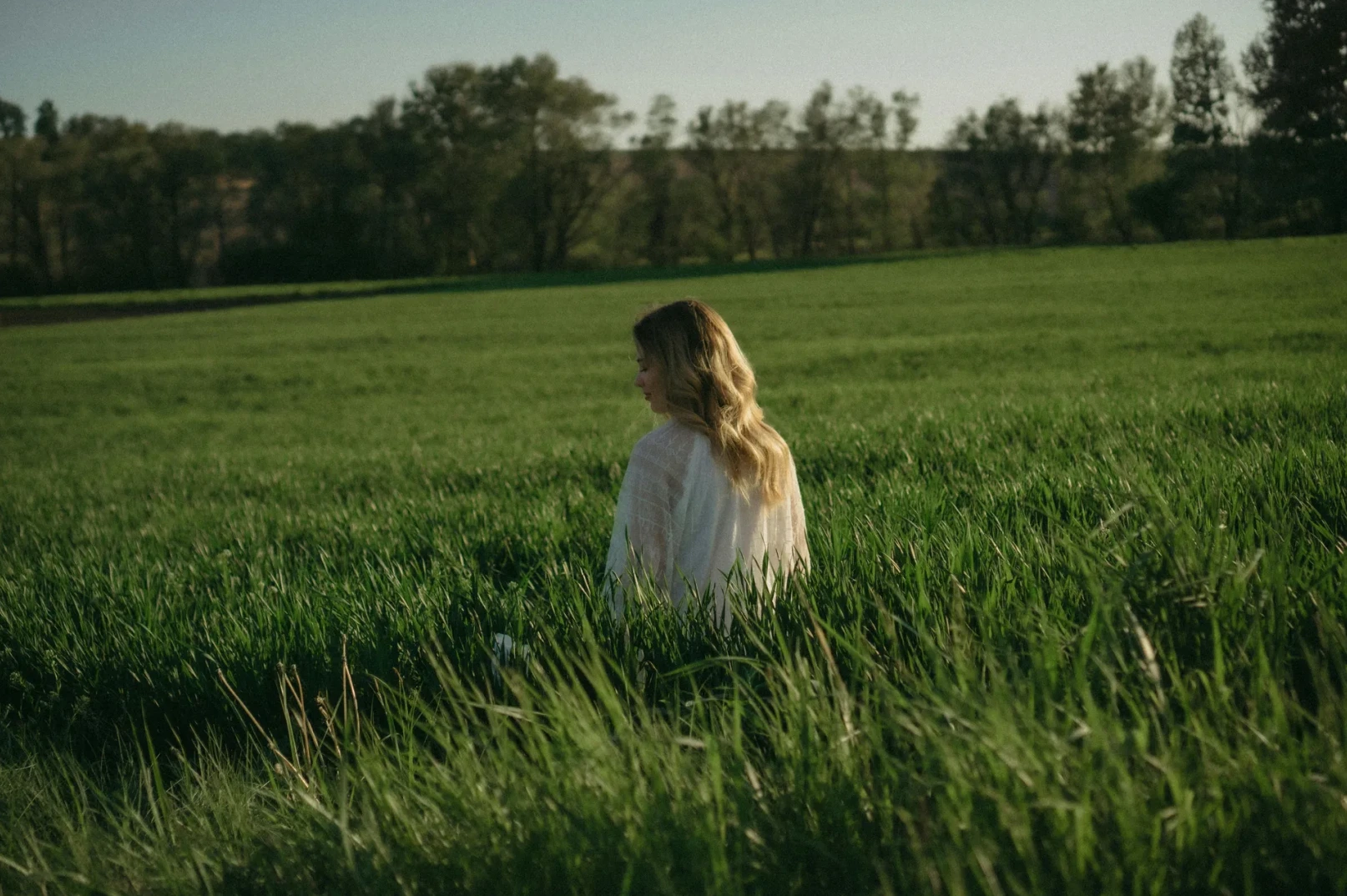 Žena sa dugom plavom kosom obučena u bijelu haljinu stoji okrenuta leđima u polju visoke zelene trave.