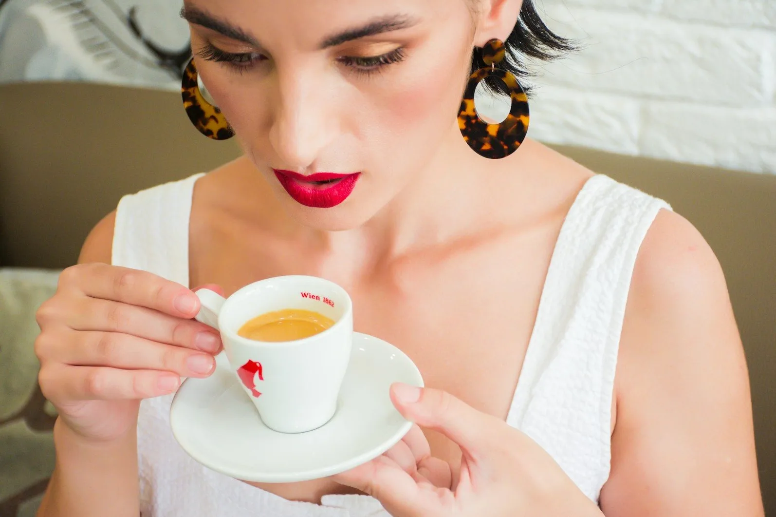 Žena s tamnom kosom i upadljivim naušnicama s uzorkom kornjačevine drži šoljicu espresa u ruci, s crvenim ružem na usnama.