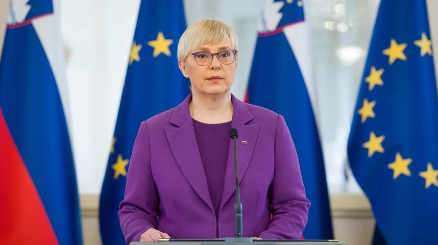 Ženska političarka nosi ljubičasto odijelo i stoji ispred zastava Slovenije i Evropske unije.