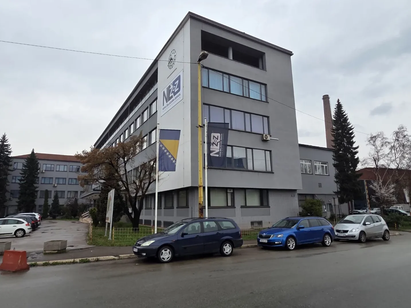 Moderna siva zgrada s natpisom "NZ" i zastavom Bosne i Hercegovine, parkirani automobili ispred.
