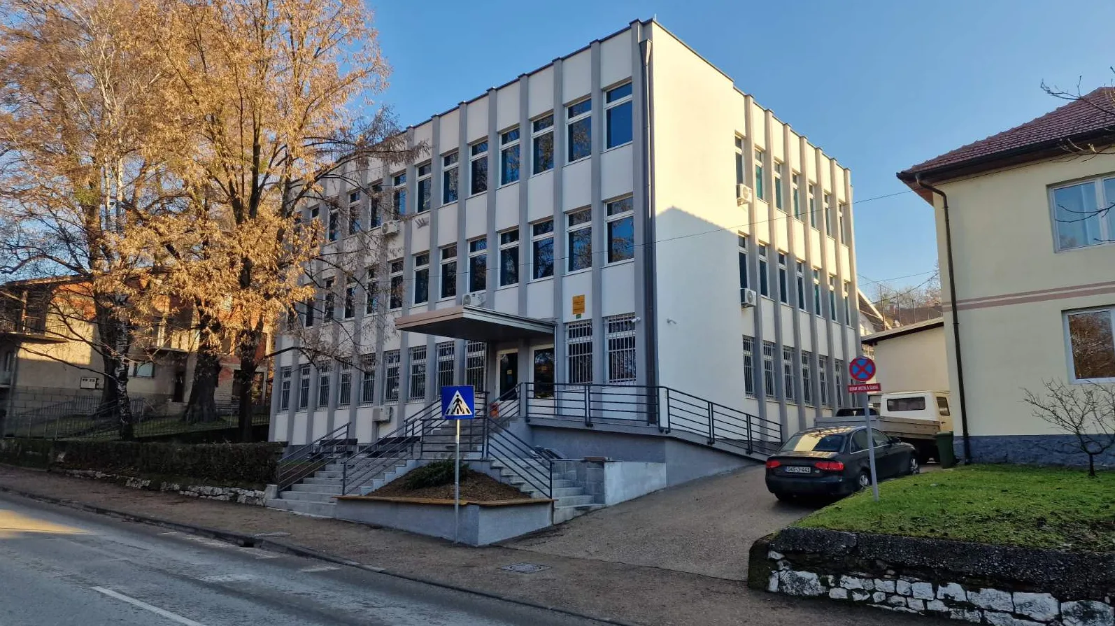 Moderna bijela zgrada javne ustanove sa mnogo prozora, stepenicama i pristupnim rampama, u jesenjem danu