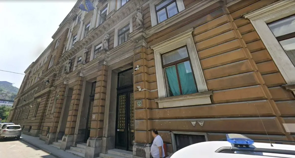 Fasada smeđe zgrade sa velikim prozorima i ukrasima, te zastavom Bosne i Hercegovine na vrhu.