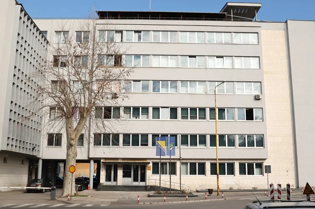Moderna siva zgrada sa velikim prozorima, ispred koje se nalazi drvo bez lišća i zastave Bosne i Hercegovine i Evropske unije.