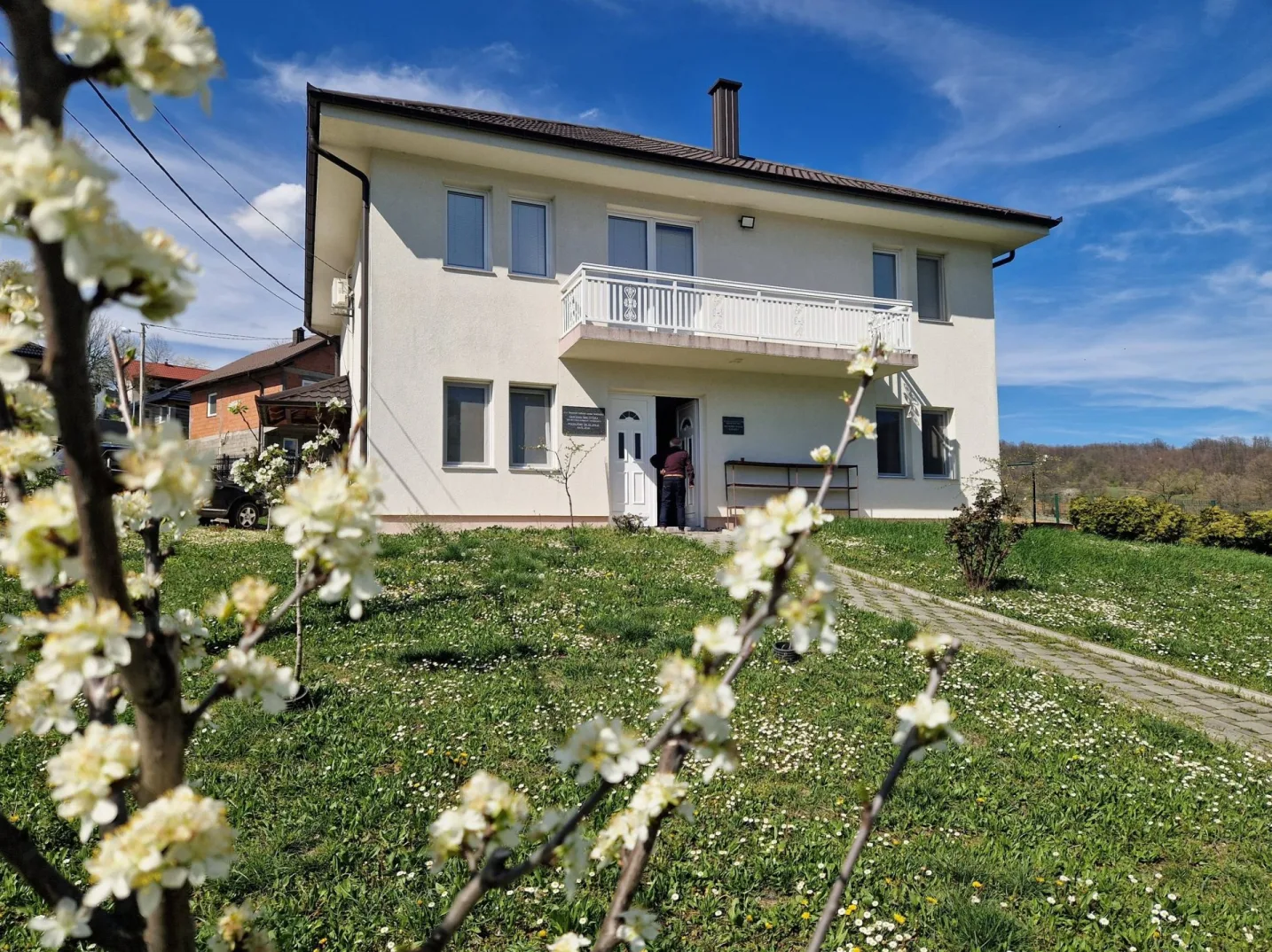 Bijela dvospratna zgrada sa bijelim balkonom i dimnjakom na krovu, okružena zelenom travom posutom cvijećem i drvećem u cvatu.