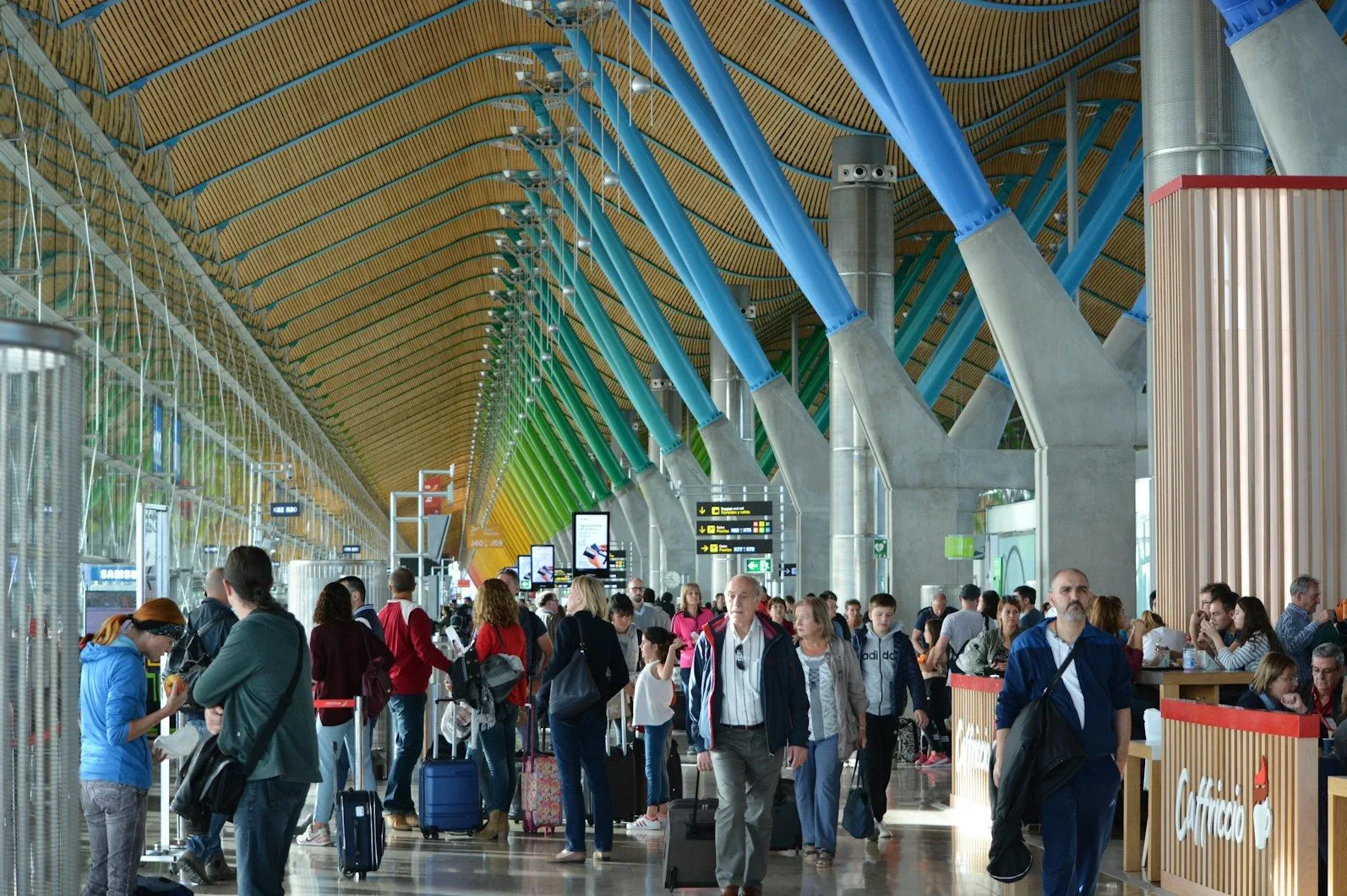 Putnici hodaju hodnikom modernog aerodroma sa karakterističnim plavim i zelenim stubovima i drvenim stropom.