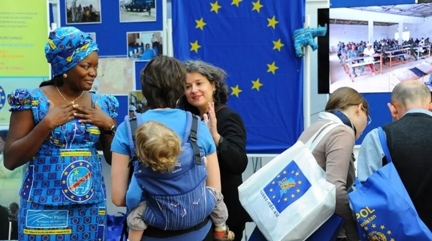 Žena u tradicionalnoj plavoj haljini sa šarom EU zastave, sa bebom u nosiljci, i drugi ljudi ispred zastave EU.