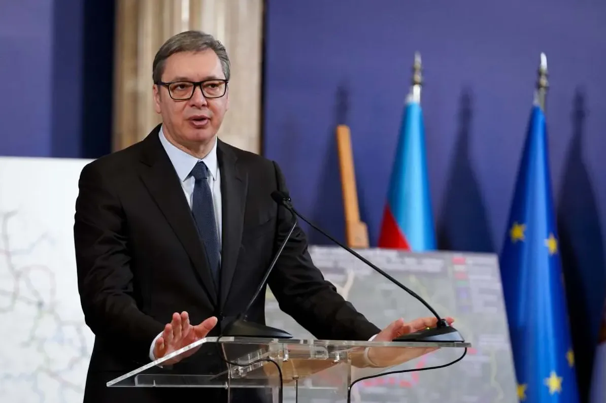 Aleksandar Vučić u crnom odijelu i plavoj kravati, s naočarima, drži govor iza prozirnog podijuma.
