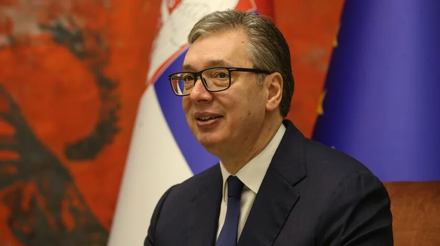 Predsjednik Srbije Aleksandar Vučić sa naočarima i tamnim odijelom, sjedi ispred zastave Srbije i Evropske unije.