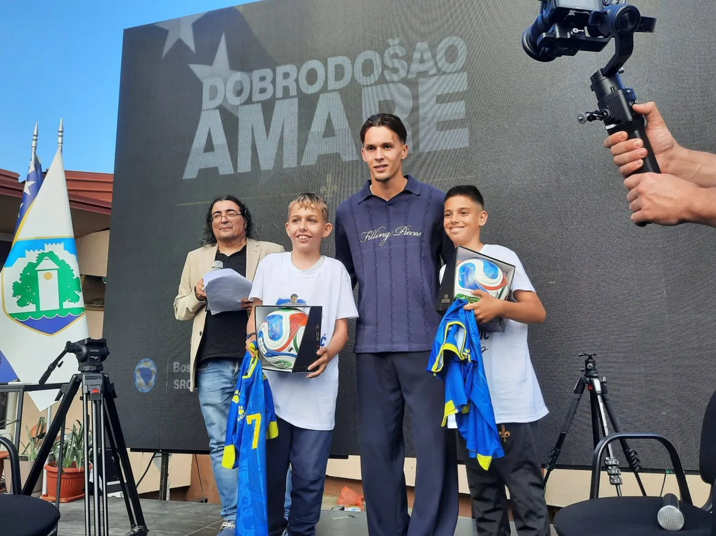 Dječaci primaju fudbalske lopte i dresove na ceremoniji dobrodošlice, pored odraslih osoba i velikog ekrana na kojem piše 'DOBRODOŠAO AMARE'.