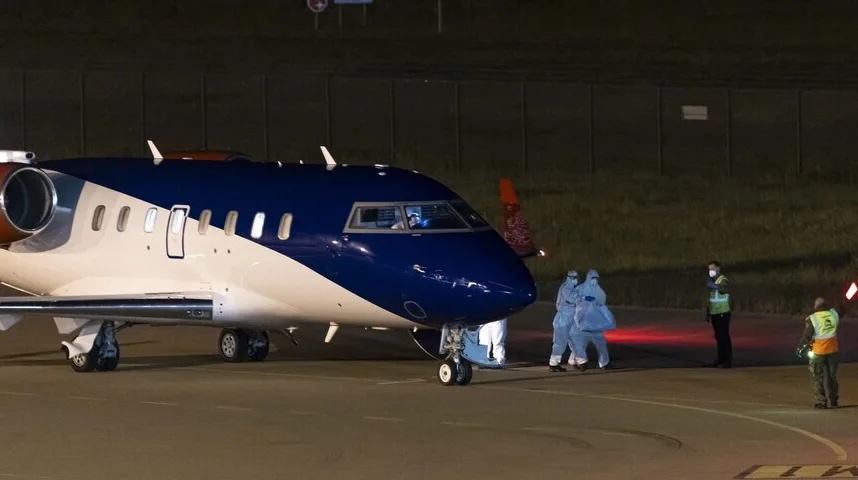 Avion sa plavo-bijelim trupom na aerodromu noću, sa osobama u zaštitnim odijelima i osobljem aerodroma na pisti.