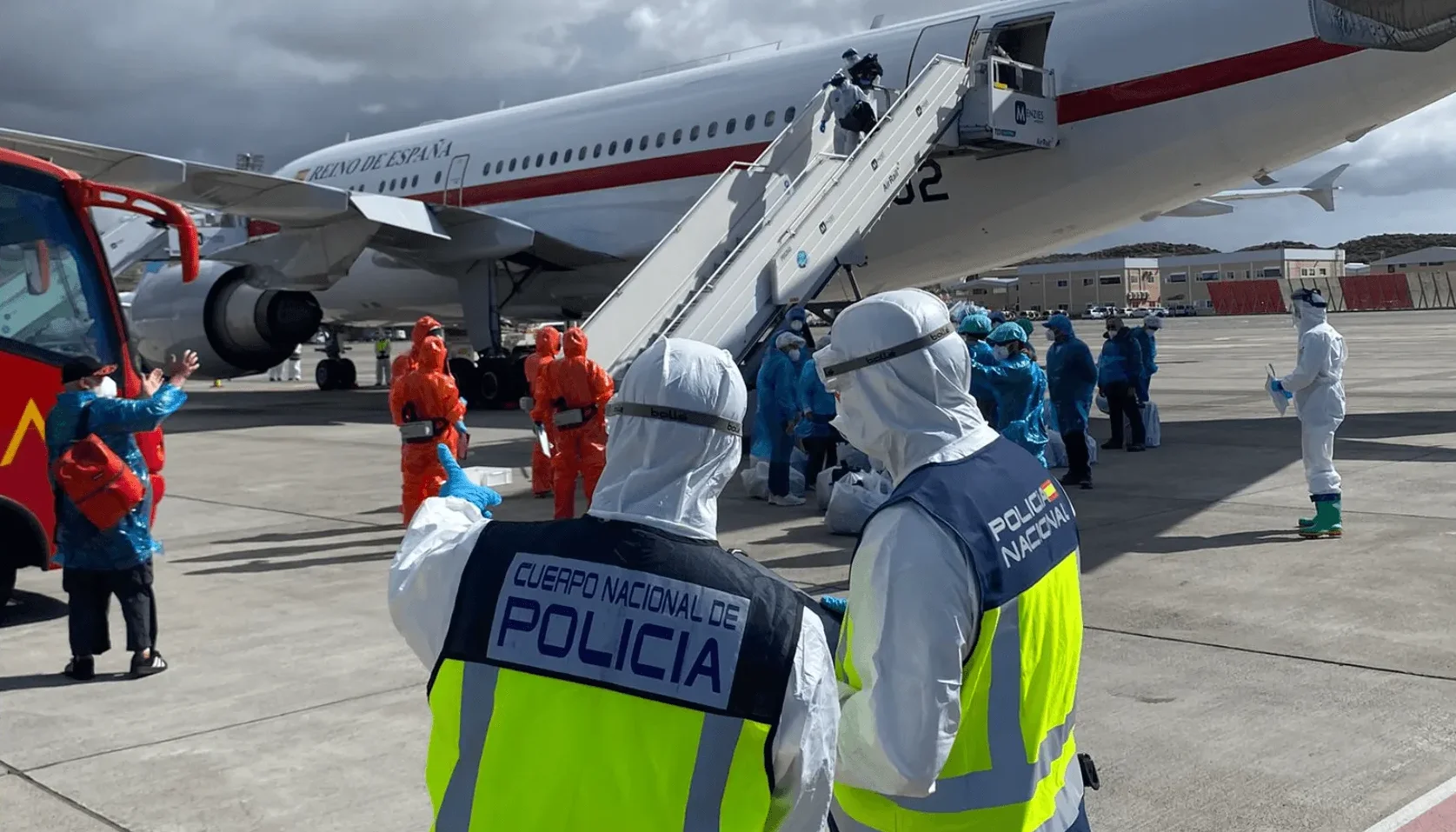 Policajci u zaštitnim odijelima na aerodromu ispred aviona Kraljevskih španskih zračnih snaga, dok se putnici ukrcavaju/iskrcavaju.