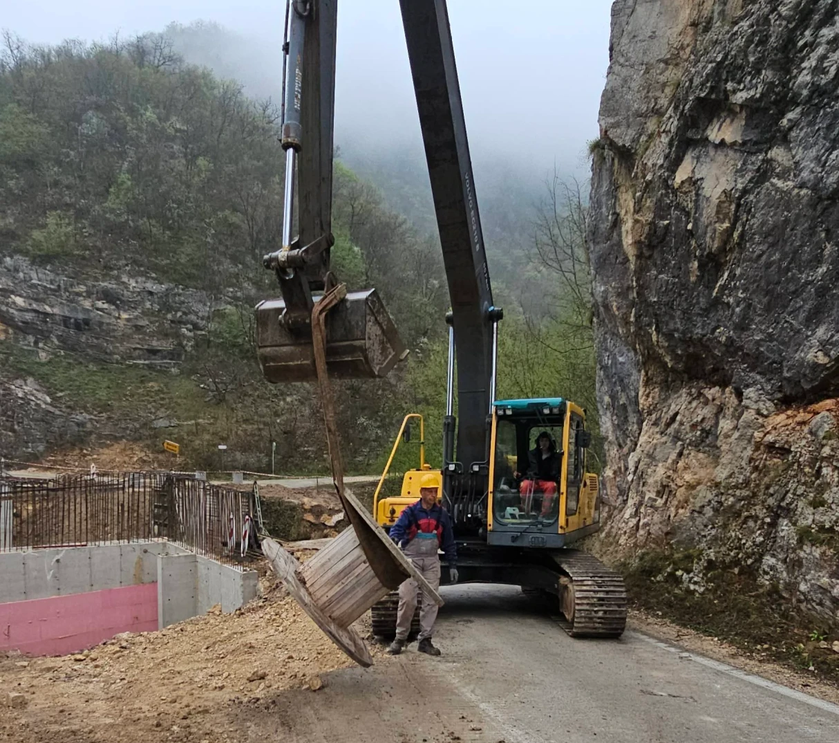 Bager sa podignutom kašikom koja drži veliku drvenu koturu, pored radnika u zaštitnoj opremi, u kanjonu.