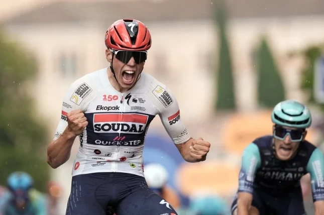 Biciklista u bijelom dresu sa crvenim i plavim detaljima, sa logom Soudal Quick-Step, proslavlja pobjedu sa podignutim pesnicama i otvorenih usta.
